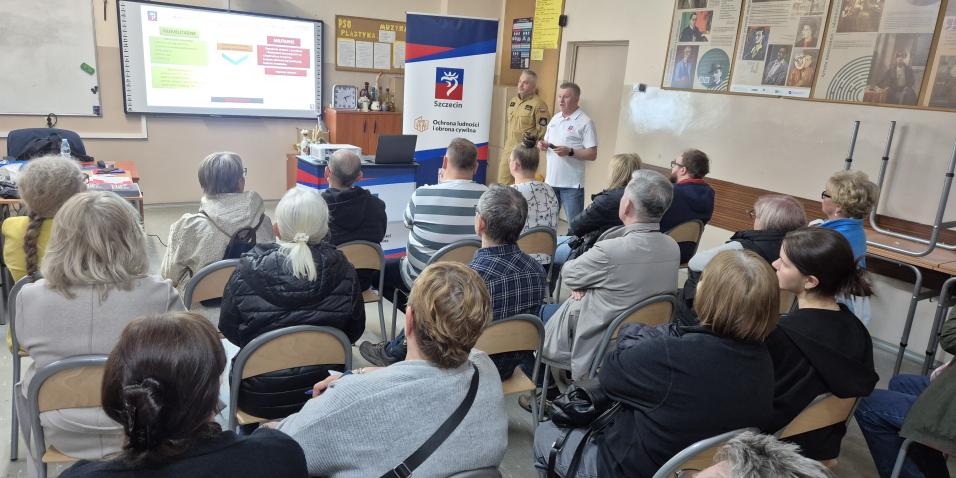 Na zdjęciu widoczna jest sala lekcyjna podczas szkolenia z zakresu ochrony ludności i obrony cywilnej organizowanego dla mieszkańców w Szkole Podstawowej nr 37 w Szczecinie. W pomieszczeniu siedzi około trzydziestu osób w różnym wieku, ustawionych na krzesłach w kilku rzędach i skierowanych w stronę prowadzących szkolenie.  Na przedzie sali znajdują się dwaj instruktorzy. Jeden z nich ubrany jest w jasną koszulkę z logo Szczecina, drugi w mundur służbowy. Obok nich ustawiony jest roll-up z herbem Szczecina 